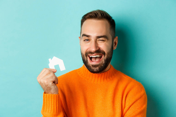 Koncept nemovitostí. Šťastný mladý muž hledá bydlení, drží domácí papír maket a usmívá se, stojí nad modrým pozadím - Fotografie, Obrázek