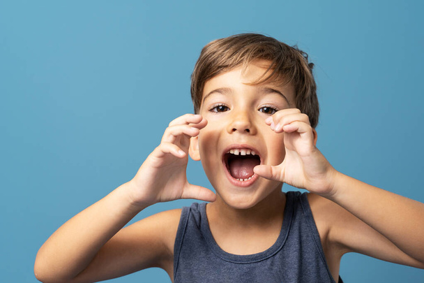 Vorderansicht eines kleinen kaukasischen Jungen von vier Jahren, der vor blauem Hintergrund vor einem Studio steht und die Hände vor seinem geöffneten Mund hält, als würde er schreien - Foto, Bild