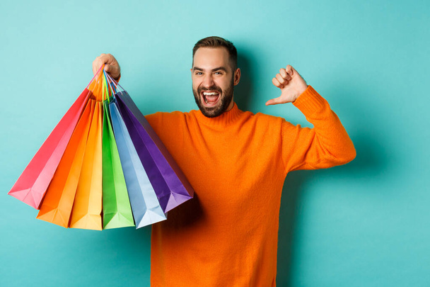 Handsome bearded man in orange sweater, buying gifts, pointing at himself and showing shopping bags, standing over blue background - Foto, Imagen