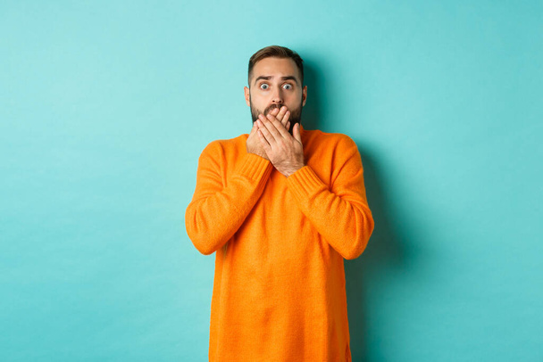 Homem atordoado olhando para cena horrível, ofegante e olhar com medo, de pé em suéter de inverno laranja, fundo do estúdio - Foto, Imagem