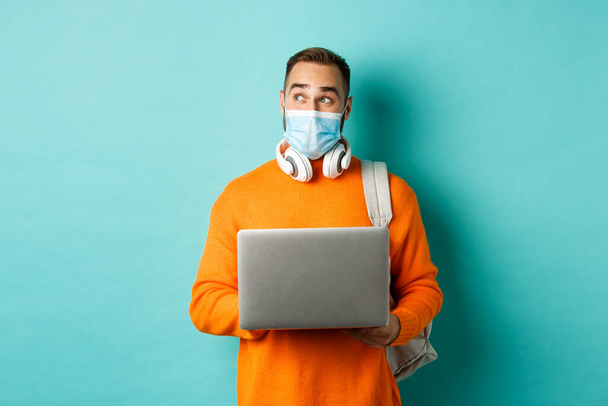 Jovem freelancer feliz em máscara médica, trabalhando no laptop, olhando para o lado pensativo, de pé sobre fundo turquesa - Foto, Imagem