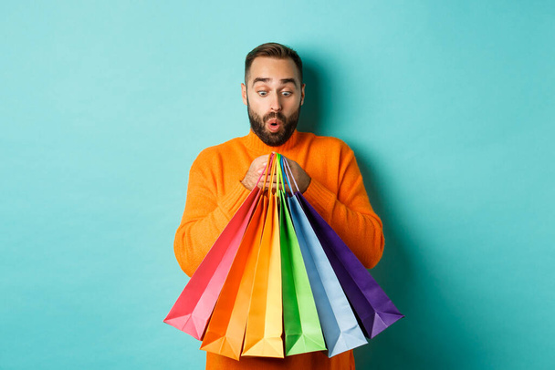 Excited man looking at shopping bags with purchases, standing amused against turquoise background - Photo, Image