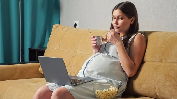 Zwangere vrouw die yoghurt en popcorn eet en naar laptop kijkt terwijl ze op de bank zit - Foto, afbeelding