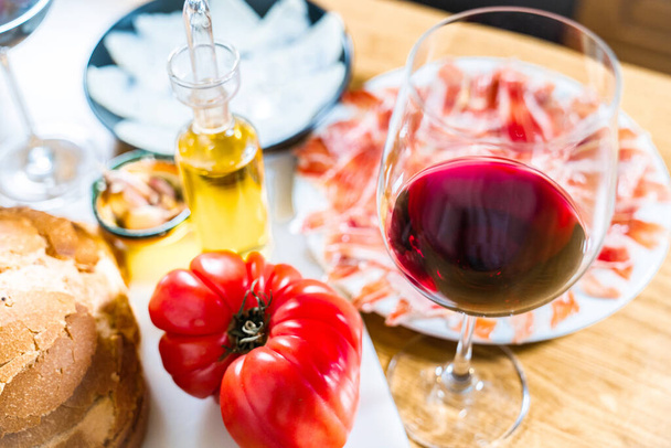 Typical Catalan or Spanish food. Bread with tomato, Iberian ham, glass of red wine, goat cheese, oil and garlic. Food products for dinner or tapas on a wooden background. - Фото, зображення