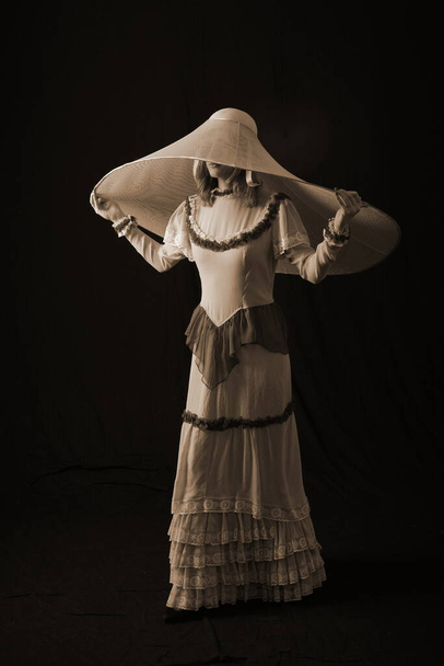 A girl in an old dress and hat stands on a dark background and is sad - Foto, imagen