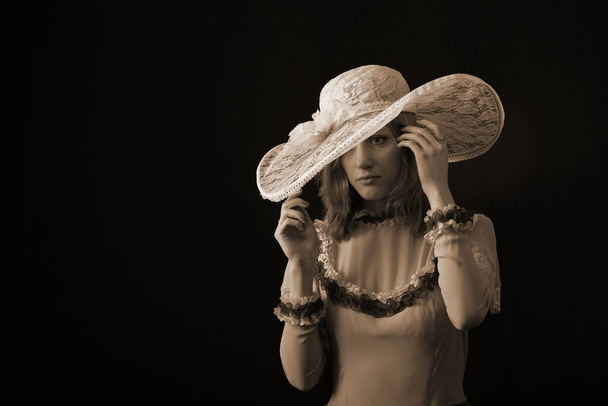 A girl in an old dress and hat stands on a dark background and is sad - Фото, изображение