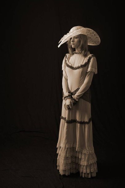 A girl in an old dress and hat stands on a dark background and is sad - Фото, изображение