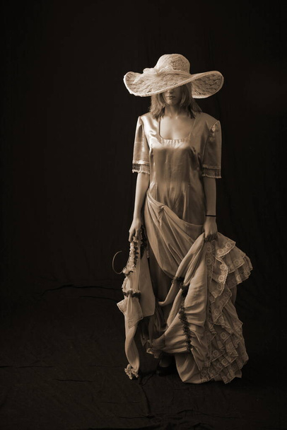 A girl in an old dress and hat stands on a dark background and is sad - Fotó, kép
