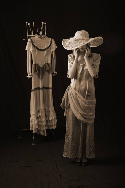 A girl in an old dress and hat stands on a dark background and is sad - 写真・画像