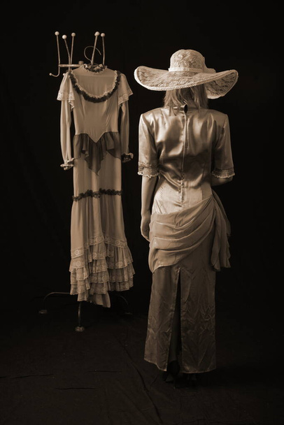 Een meisje in een oude jurk en hoed staat op een donkere achtergrond en is verdrietig - Foto, afbeelding