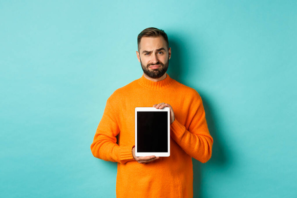 Τεχνολογία. Σκεπτικός και δυσαρεστημένος τύπος που δείχνει ψηφιακή οθόνη tablet, συνοφρυωμένος απογοητευμένος, στέκεται πάνω από το γαλάζιο φόντο - Φωτογραφία, εικόνα