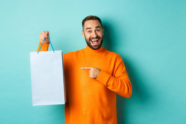 Όμορφος χαμογελαστός άντρας που δείχνει με το δάχτυλο την τσάντα για ψώνια, αγοράζει σε καταστήματα, στέκεται ικανοποιημένος πάνω από το μπλε φόντο - Φωτογραφία, εικόνα