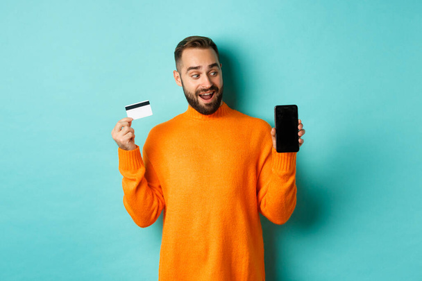 Интернет-магазины. Удивительный парень с помощью кредитной карты и показывая мобильный экран, глядя впечатлен, стоя на светло-голубом фоне - Фото, изображение