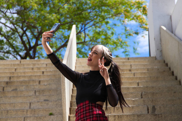 ładna młoda dziewczyna w punkowym stylu robi zdjęcia telefonem komórkowym na schodach w parku. Koncepcja technologii i piękna - Zdjęcie, obraz