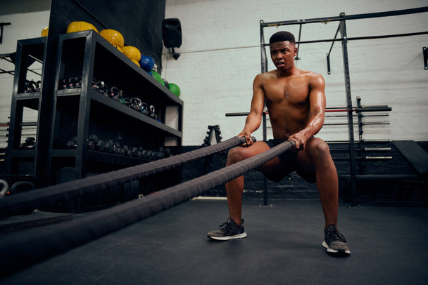 Young African American male exercising inside a gym. Topless male personal trainer using rope to strengthen his arms. High quality photo - Foto, afbeelding