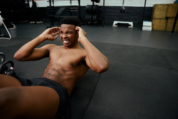 Young African American male doing sit-ups on the floor in the gym. Mixed race male personal trainer exercising shirtless indoors. High quality photo - Fotó, kép