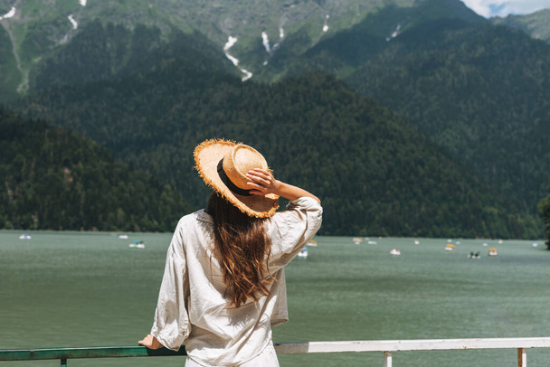 Νεαρή γυναίκα ταξιδιώτη με μακριά ξανθά μαλλιά σε ψάθινο καπέλο κοιτάζει όμορφη θέα της λίμνης βουνό, οι άνθρωποι από πίσω - Φωτογραφία, εικόνα