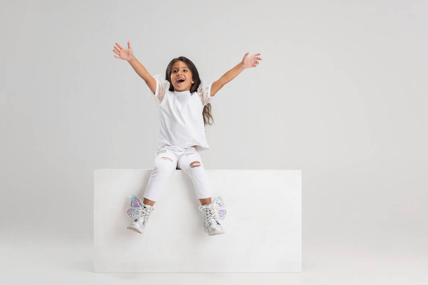 Portret van een klein schattig blank meisje dat op een grote doos zit, geïsoleerd boven een witte studio achtergrond. Ziet er gelukkig uit, vrolijk - Foto, afbeelding