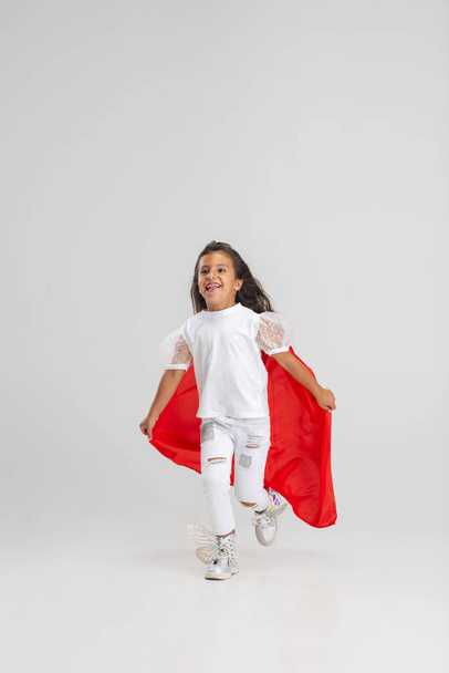 Small superhero. Little cute Caucasian girl in a red cape isolated over white studio background. Full length portrait - Foto, afbeelding