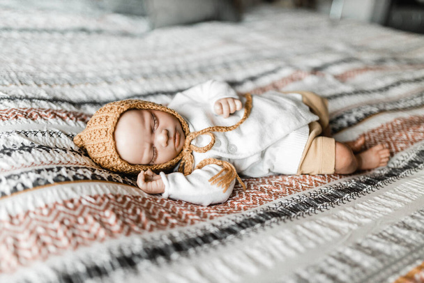 Reborn baby doll lying on a bed with a patterned dovet cover wearing a bonnet, model release not required as baby is a lifelike doll - Photo, Image