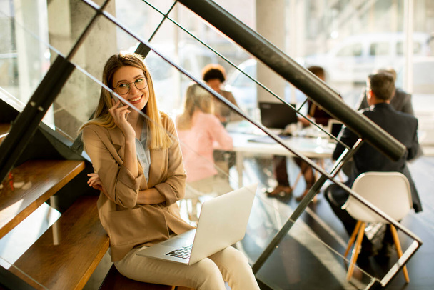 Красива молода жінка сидить на сходах з ноутбуком і використовує мобільний телефон в сучасному офісі перед своєю командою
 - Фото, зображення
