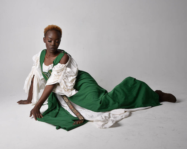 Ganztägiges Porträt einer hübschen afrikanischen Frau, die ein langes grünes mittelalterliches Fantasiegewand trägt und auf hellgrauem Studiohintergrund sitzt. - Foto, Bild