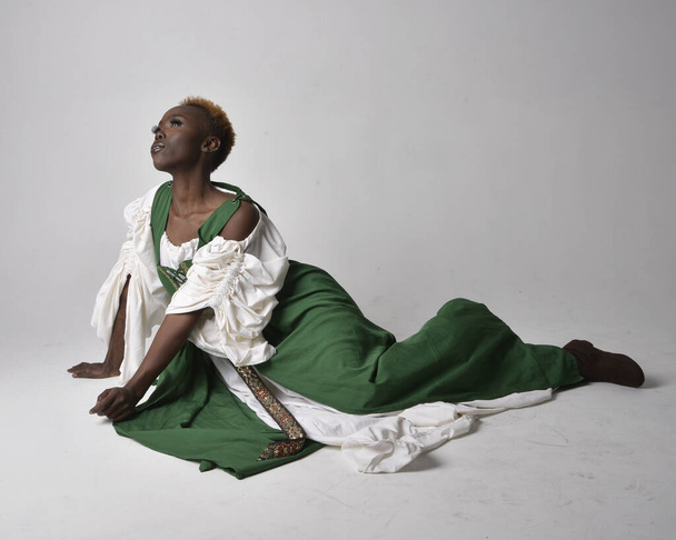Full length portrait of pretty African woman wearing long green medieval fantasy gown, sitting on a light grey studio background. - Φωτογραφία, εικόνα