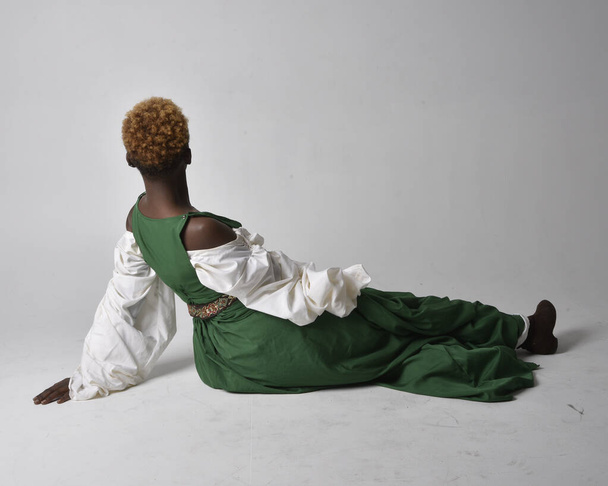 Full length portrait of pretty African woman wearing long green medieval fantasy gown, sitting on a light grey studio background. - Zdjęcie, obraz