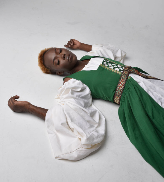 Full length portrait of pretty African woman wearing long green medieval fantasy gown, sitting on a light grey studio background. - Photo, Image