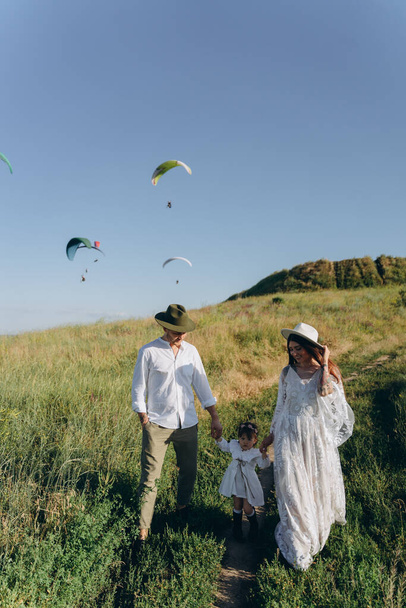 beautiful woman in white guipure dress with her husband and daughter walking in the field - Фото, зображення