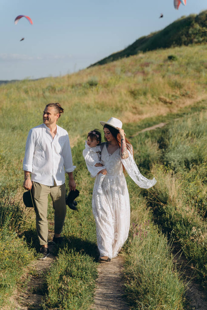 beautiful woman in white guipure dress holding her daughter and walking with husband in the field - Foto, imagen