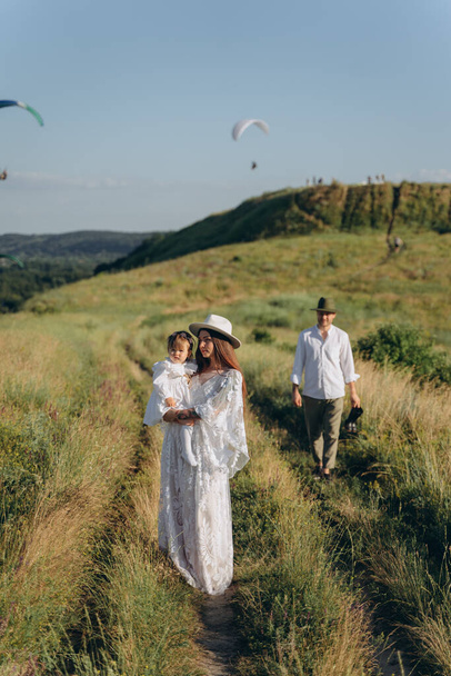 beautiful woman in white guipure dress holding her daughter and walking with husband in the field - Valokuva, kuva