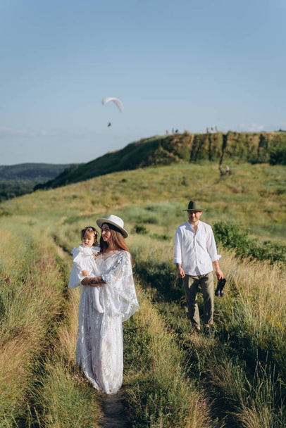 beautiful woman in white guipure dress holding her daughter and walking with husband in the field - Foto, Bild