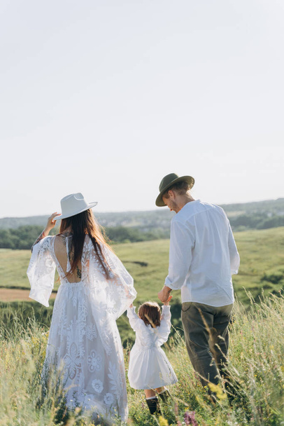 Happy young family spending  time together in nature, beautiful woman in white guipure dress with husband and their daughter walking  in the field - Photo, Image