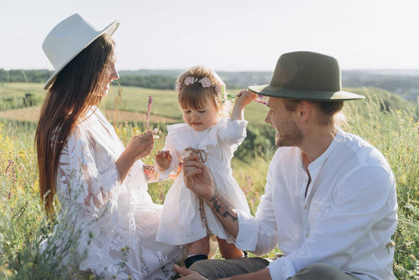 Feliz familia joven pasar tiempo juntos en la naturaleza, hermosa mujer en vestido de guipur blanco con el marido y su hija sentada en el campo - Foto, imagen