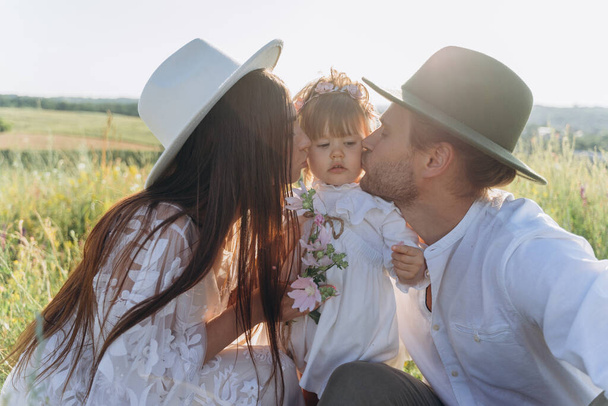 Happy young family spending  time together in nature, beautiful woman in white guipure dress with husband kissing their daughter  - Фото, изображение