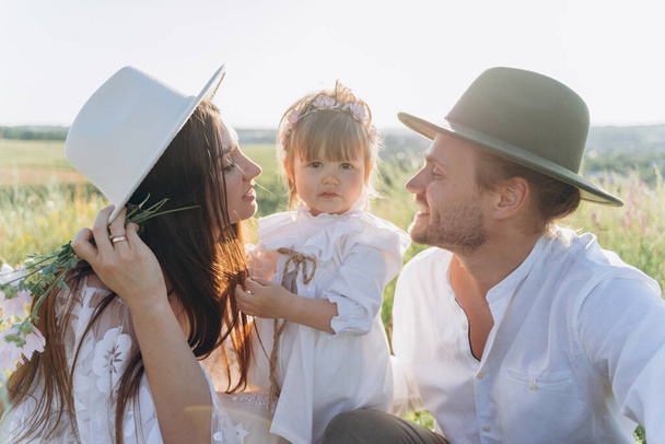 Happy young family spending  time together in nature, beautiful woman in white guipure dress with husband and their daughter sitting in the field - Foto, Imagen