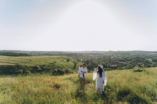 Happy young family spending  time together in nature, beautiful woman in white guipure dress with husband and their daughter walking  in the field - 写真・画像