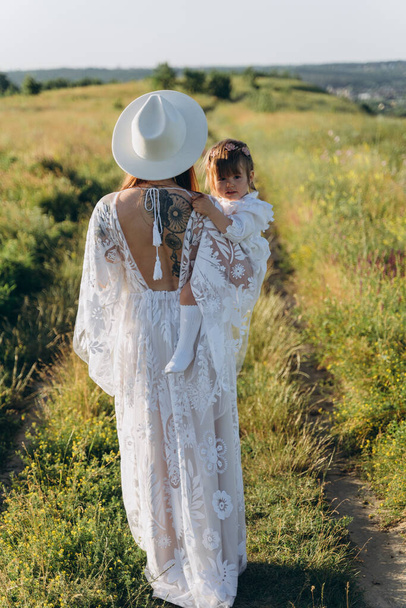 La hermosa mujer en vestido de guipur blanco sosteniendo a su hija y caminando en el campo - Foto, Imagen