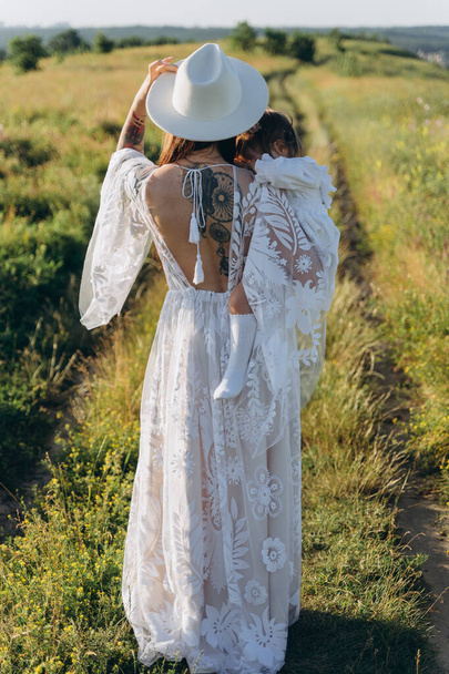 Красива жінка в білій сукні-гіпюрі тримає свою дочку і ходить у полі
 - Фото, зображення