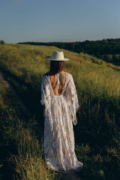 The beautiful woman in white guipure dress and in hat posing in the field, standing with her back to the camera - Foto, imagen