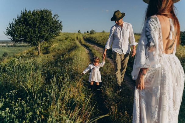 Happy young family spending  time together in nature, beautiful woman in white guipure dress with husband and their daughter walking  in the field - Fotó, kép
