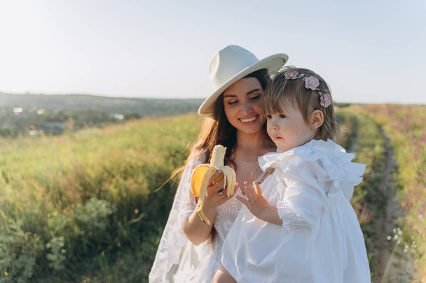 The beautiful woman in white guipure dress holding her daughter and eating a banana in the field - Fotoğraf, Görsel