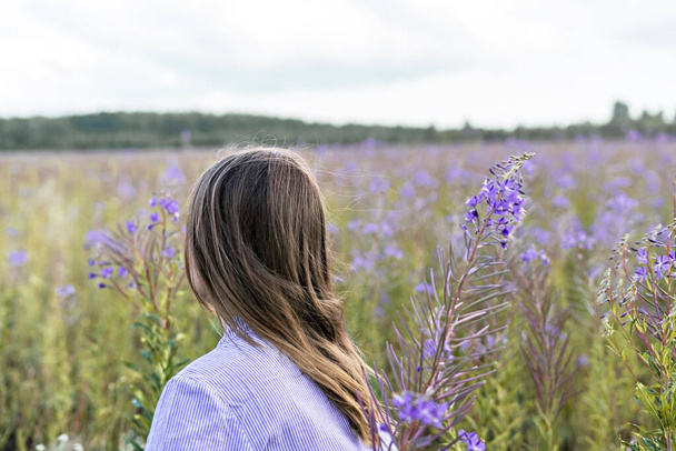 Young beautiful blond woman in purple shirt from behind walking in the meadow among flowers of fireweed, beauty in nature landscape selective focus - Photo, Image