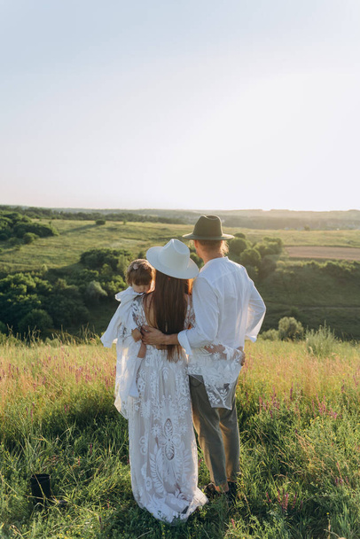 Happy young family spending  time together in nature, beautiful woman in white guipure dress holding her daughter and walking with husband in the field - Φωτογραφία, εικόνα