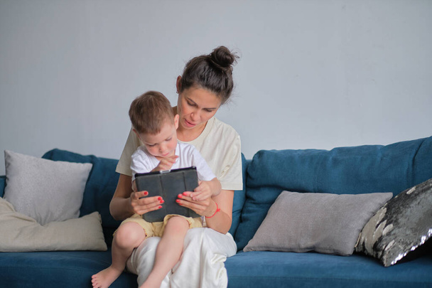 家にタブレットを持ってる母親と息子。家のインテリアのリビングルームのソファの上に座っているタブレットで彼女の息子にオンラインでメディアコンテンツを示す母 - 写真・画像