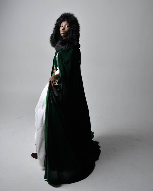 Portret pięknej afrykańskiej kobiety noszącej długą zieloną średniowieczną suknię fantazyjną i aksamitną pelerynę, siedzącej na jasnoszarym tle studia. - Zdjęcie, obraz