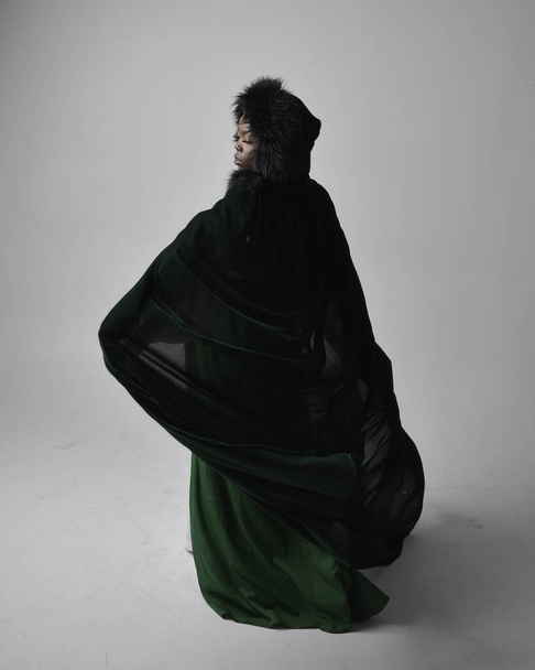 Full length portret van mooie Afrikaanse vrouw met lange groene middeleeuwse fantasie jurk en fluwelen mantel, zittend op een lichtgrijze studio achtergrond. - Foto, afbeelding