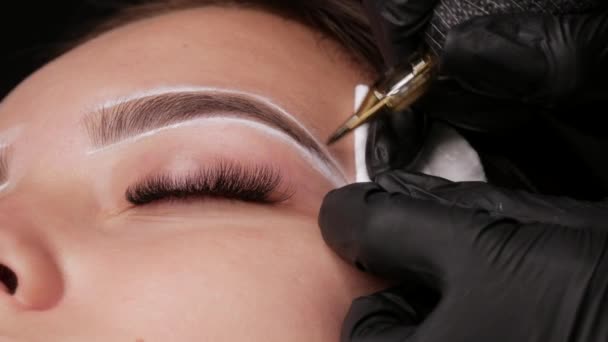 Master applies dark coloring pigment to correct shape of eyebrows with a special machine. The beautiful face of young girl model on sofa in a beauty salon before microblading, permanent makeup tattoo - Footage, Video