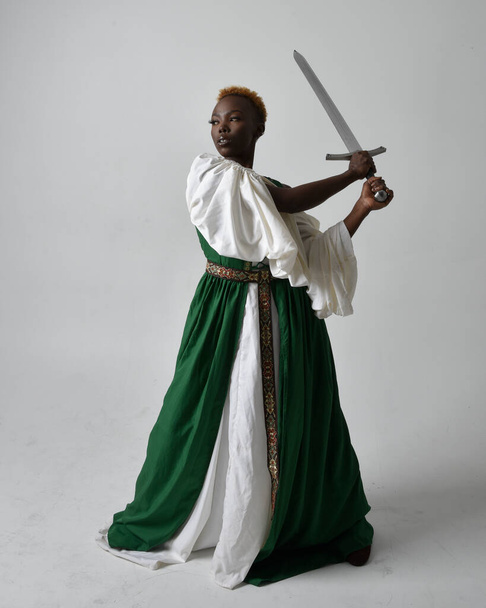 Pełna długość portret pięknej afrykańskiej kobiety noszącej długą zieloną średniowieczną suknię fantasy trzymającej miecz, stojącej akcji poza na jasnoszarym tle studio. - Zdjęcie, obraz
